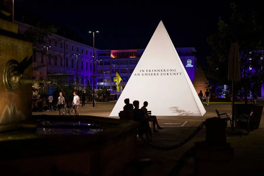 Die beleuchtete Pyramide am Abend, auf der Fläche steht „In Erinnerung an unsere Zukunft“